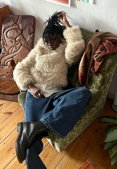 Chaqueta de piel sintética en color crema, pantalones anchos de rayas azules, botas negras con cordones. Sentada en una silla verde con un patrón, con un textil estampado drapeado cerca.