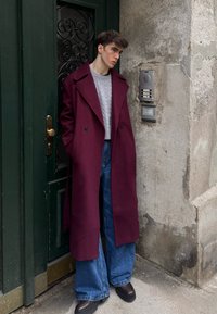 Manteau en laine bordeaux avec larges revers, associé à un pull gris à torsades, un jean bleu ample et des chaussures à plateforme foncées. Fond de porte verte.