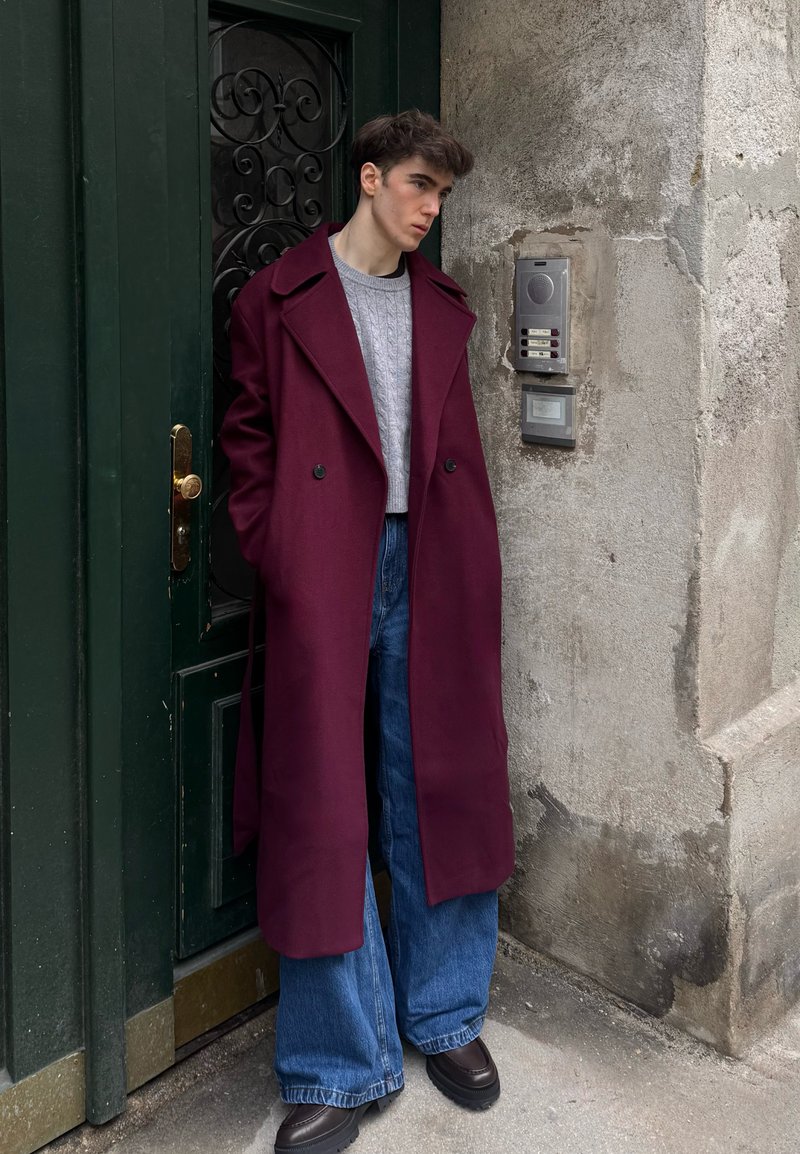 Manteau en laine bordeaux à larges revers, associé à un pull en maille torsadée gris, un jean bleu ample et des chaussures à plateforme foncées. Fond de porte verte.