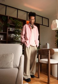 Camicetta rosa chiaro a bottoni, pantaloni beige a gamba larga, scarpe nere slip-on, sciarpa decorata e grandi occhiali rotondi in un ambiente interno moderno.