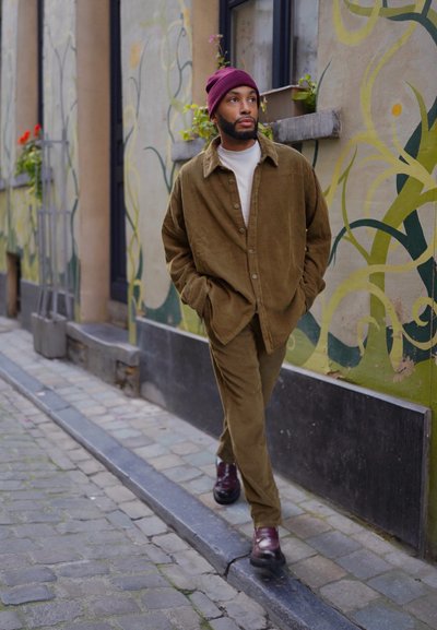 Camisa y pantalones de corduroy oliva combinados con un cuello de tortuga claro. Gorro burdeos, botas marrón oscuro, caminando por una calle adoquinada junto a un mural.