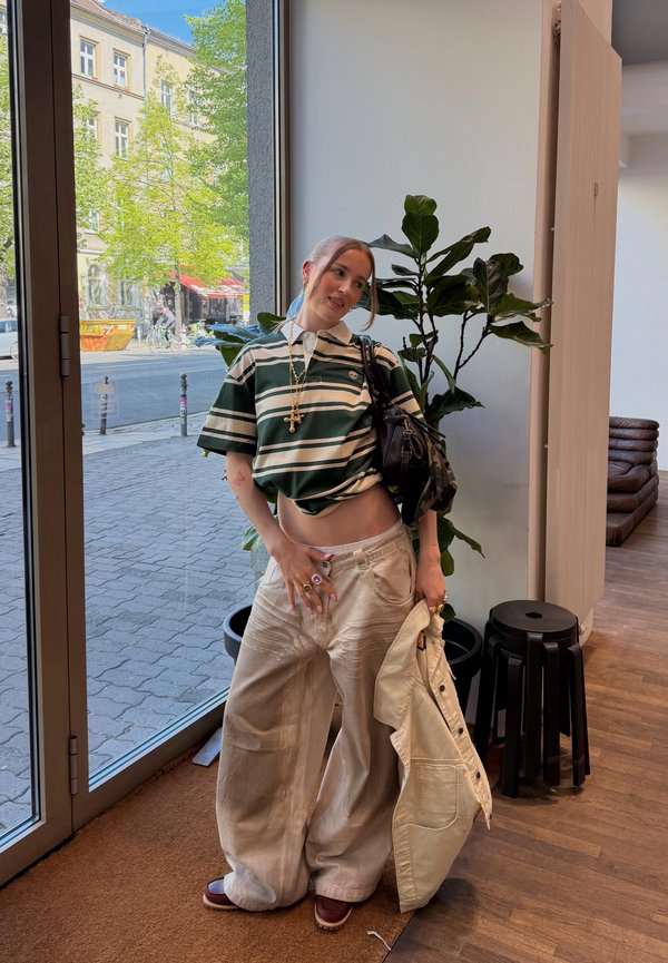 Young person in loose beige pants and striped green shirt stands indoors by glass door, holding beige jacket with potted plant in background.