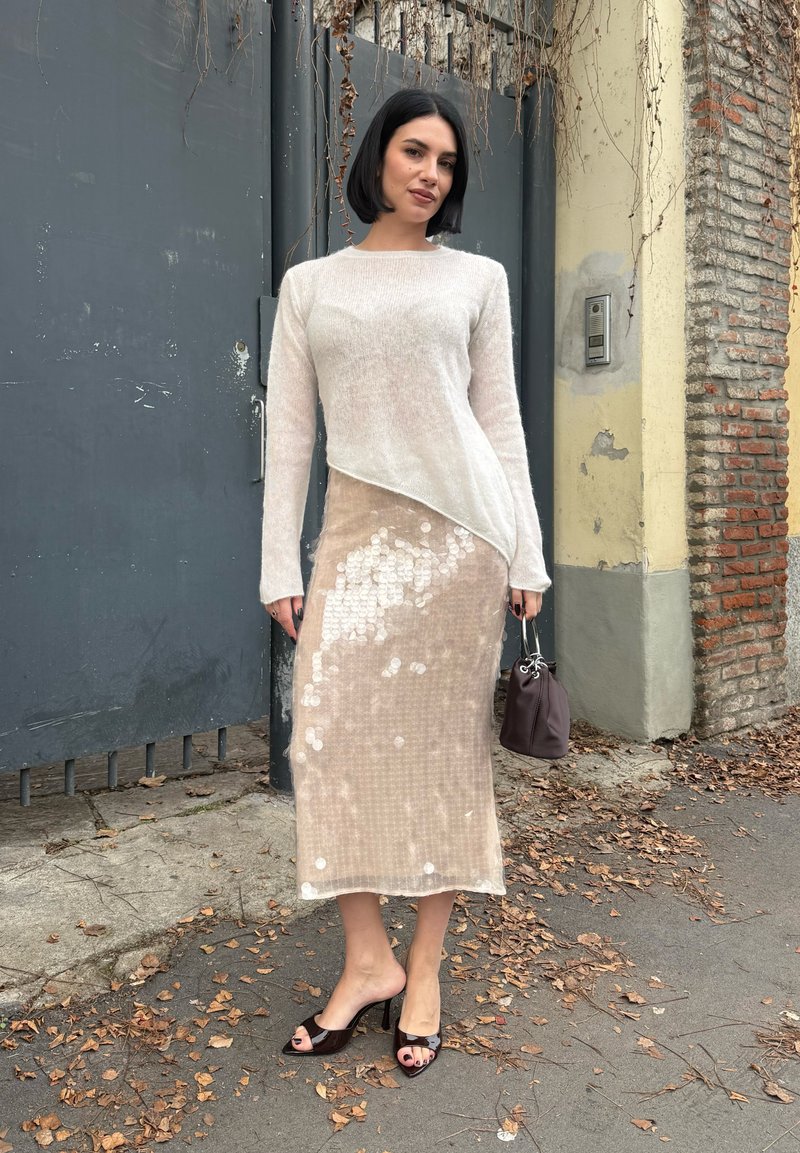 Femme aux cheveux noirs coupe au carré portant un pull beige clair asymétrique, une jupe ornée de sequins, des sandales à talons noires et tenant un petit sac violet.