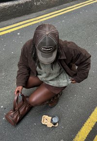 Άτομο με καφέ μπουφάν, σορτς και μπότες γονατισμένο στο δρόμο, κρατώντας μια καφέ τσάντα, κοντά σε μια ποτήρι καφέ για πακέτο μέσα σε χάρτινο θήκη.