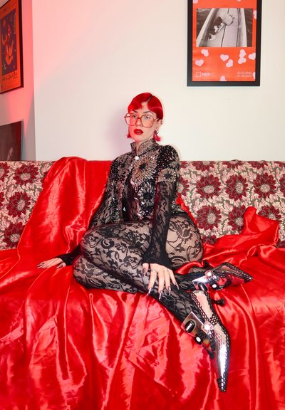 Body de encaje negro con detalles transparentes, cuello adornado; labios rojos, cabello rojo; botas de tobillo patentadas negras con acentos plateados y hebillas.