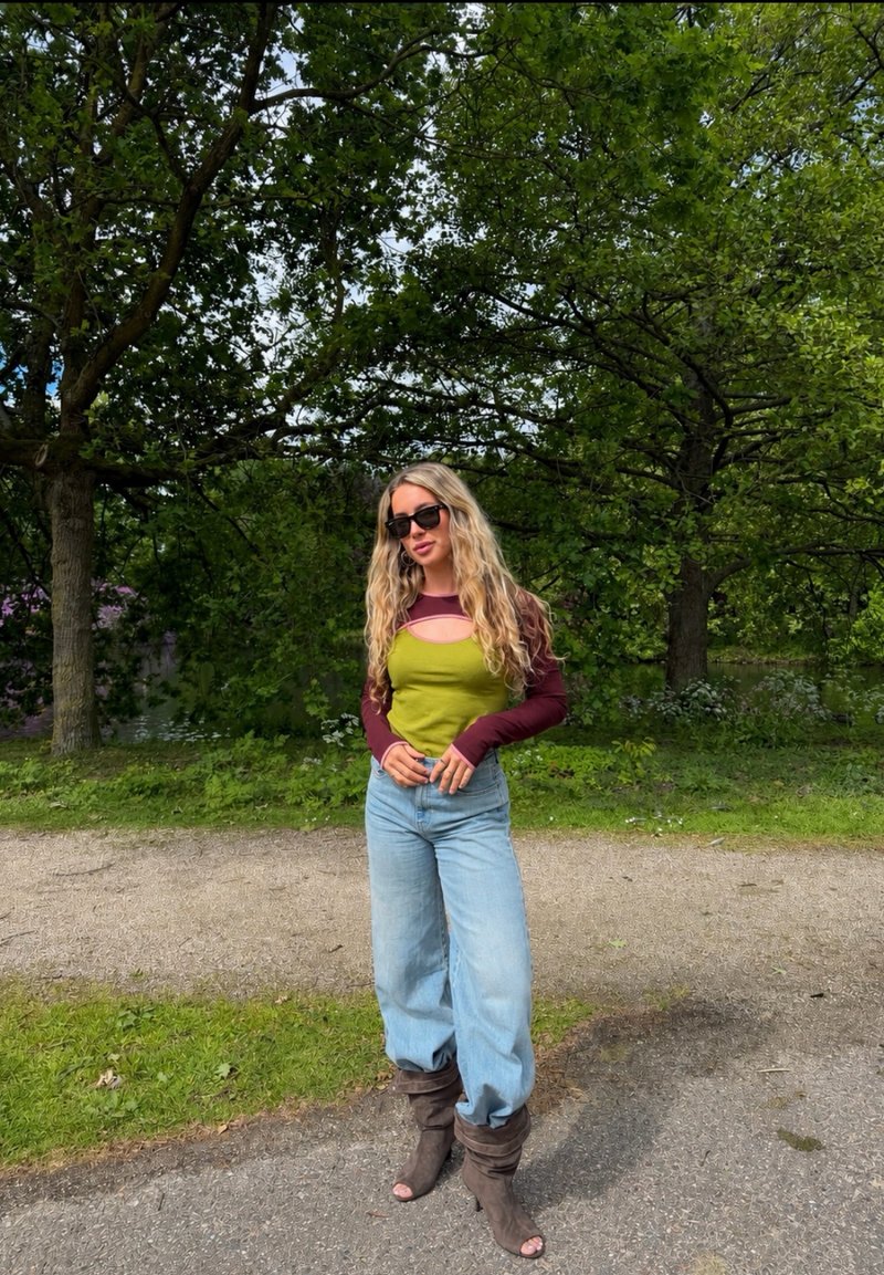 Kvinne med langt blond hår iført solbriller, grønn og burgunder topp, vide jeans og brune hellede støvler står på en sti omgitt av trær.