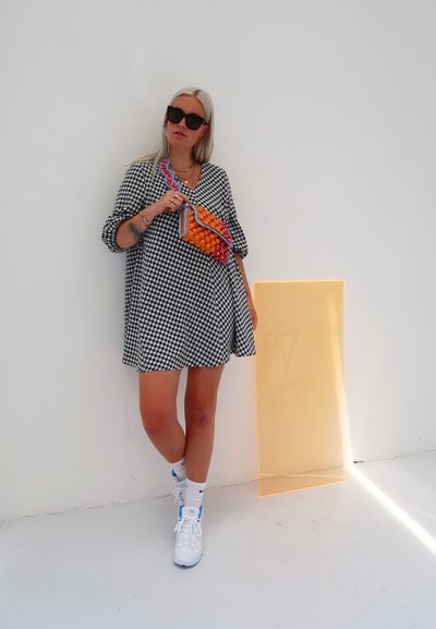 Un vestido a cuadros blanco y negro, un bolso con estampado naranja y azul, zapatillas blancas y gafas de sol. Panel amarillo transparente en el fondo.