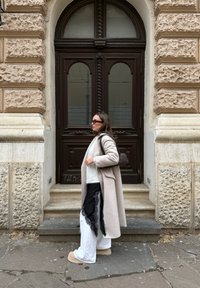 Femme portant un long manteau beige, un pantalon blanc avec une superposition en dentelle noire, des bottes beiges et des lunettes de soleil, marchant devant un bâtiment en pierre texturée avec une grande porte en bois.