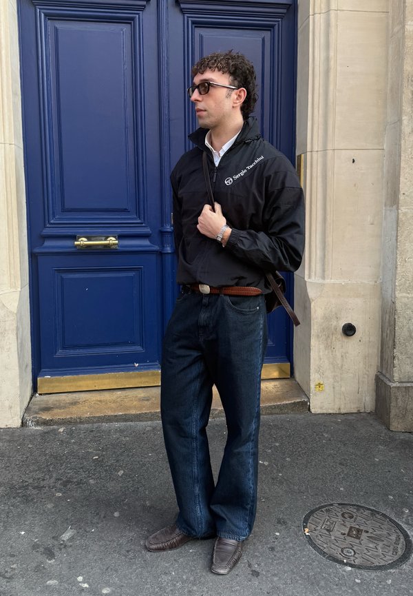 Man wearing black jacket, dark jeans, sunglasses, and brown shoes stands in front of a large blue double door on city street.