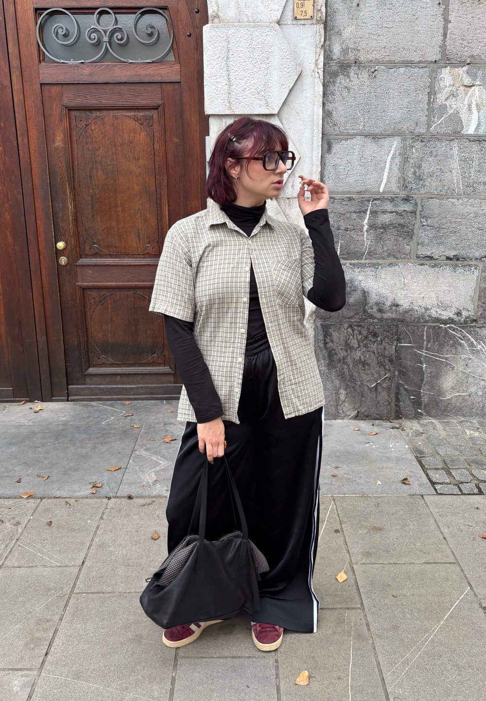 Lehká kostkovaná košile s krátkým rukávem, černé tričko s dlouhým rukávem pod ní, černé kalhoty s bílými pruhy na bocích, nese černou tašku, stojí na chodníku.