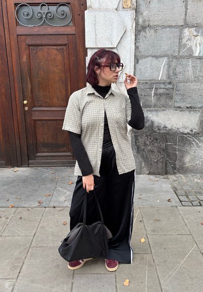 Camisa de cuadros clara de manga corta, camiseta negra de manga larga debajo, pantalones negros con rayas blancas en los lados, llevando una bolsa negra, de pie sobre el pavimento.