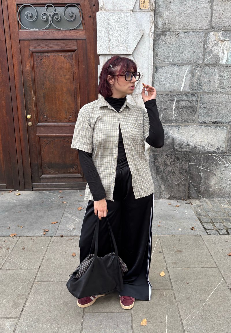 Lahka karo s kratkimi rokavi v karo vzorcu, pod njo črna majica z dolgimi rokavi, črne hlače z belo črtastimi stranicami, nosi črno torbo, stoji na tlakovcih.