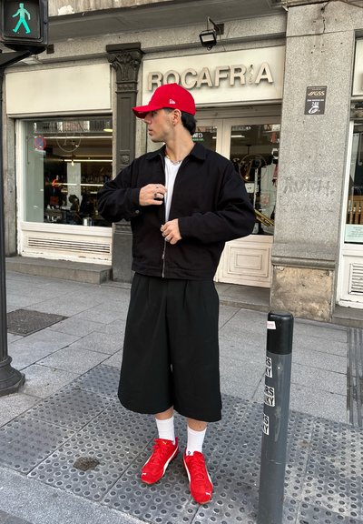 Chaqueta negra con cremallera sobre una camiseta blanca, pantalones cortos negros holgados y zapatillas rojas con calcetines blancos. Gorro rojo y joyas de plata visibles.