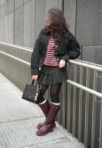 Femme en veste en jean noire, chemise rayée, jupe plissée à fines rayures, collants noirs, bottines bordeaux, tenant un sac à main noir, appuyée sur une rambarde en métal.