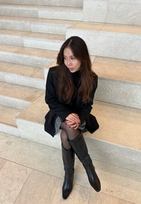 Μαύρο σακάκι πάνω από έναν κολλάρο, γκρι φούστα, μαύρες μπότες μέχρι το γόνατο. Μακριά καστανά μαλλιά, καθισμένη σε ανοιχτόχρωμες πέτρινες σκάλες, με τα χέρια να ακουμπούν στα γόνατα.