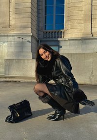 Manteau en cuir noir avec accents en fausse fourrure, bottes noires à talons hauts, collants noirs transparents, et un sac à main noir avec des ferrures dorées.