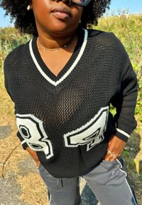 Melnā adīta džemperī ir brīvs piegriezums, baltas akcenti un lieli skaitļi "84" priekšpusē. V-veida kakla dizains ar ribotām roka daļām.