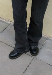 Sabots noirs en cuir à bout rond, coutures jaunes contrastantes, portés avec un pantalon évasé en denim foncé sur une surface carrelée.