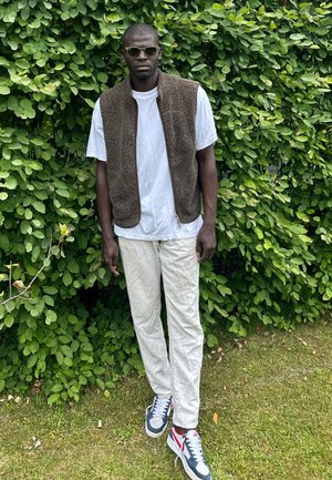 Bruine fleece vest over een wit T-shirt, gecombineerd met lichtgekleurde broek en blauwe sneakers met rode accenten. Groene, loofachtige achtergrond.