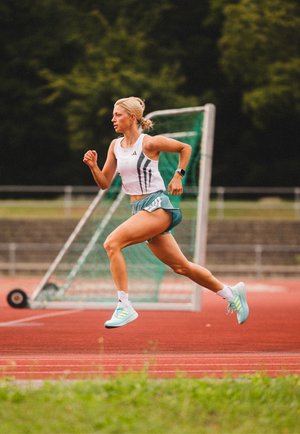 Una corredora con una camiseta sin mangas blanca con rayas verdes y pantalones cortos azul claro. Lleva zapatillas deportivas azul claro a juego en una pista roja.