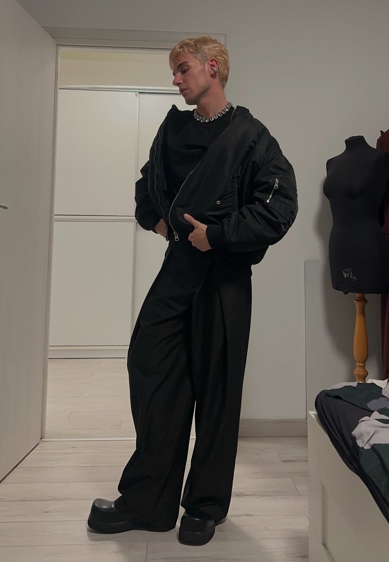 Chaqueta bomber negra con cremalleras, pantalones anchos negros y zapatos de plataforma negros y gruesos. Ambiente de habitación con muebles blancos y una maniquí.