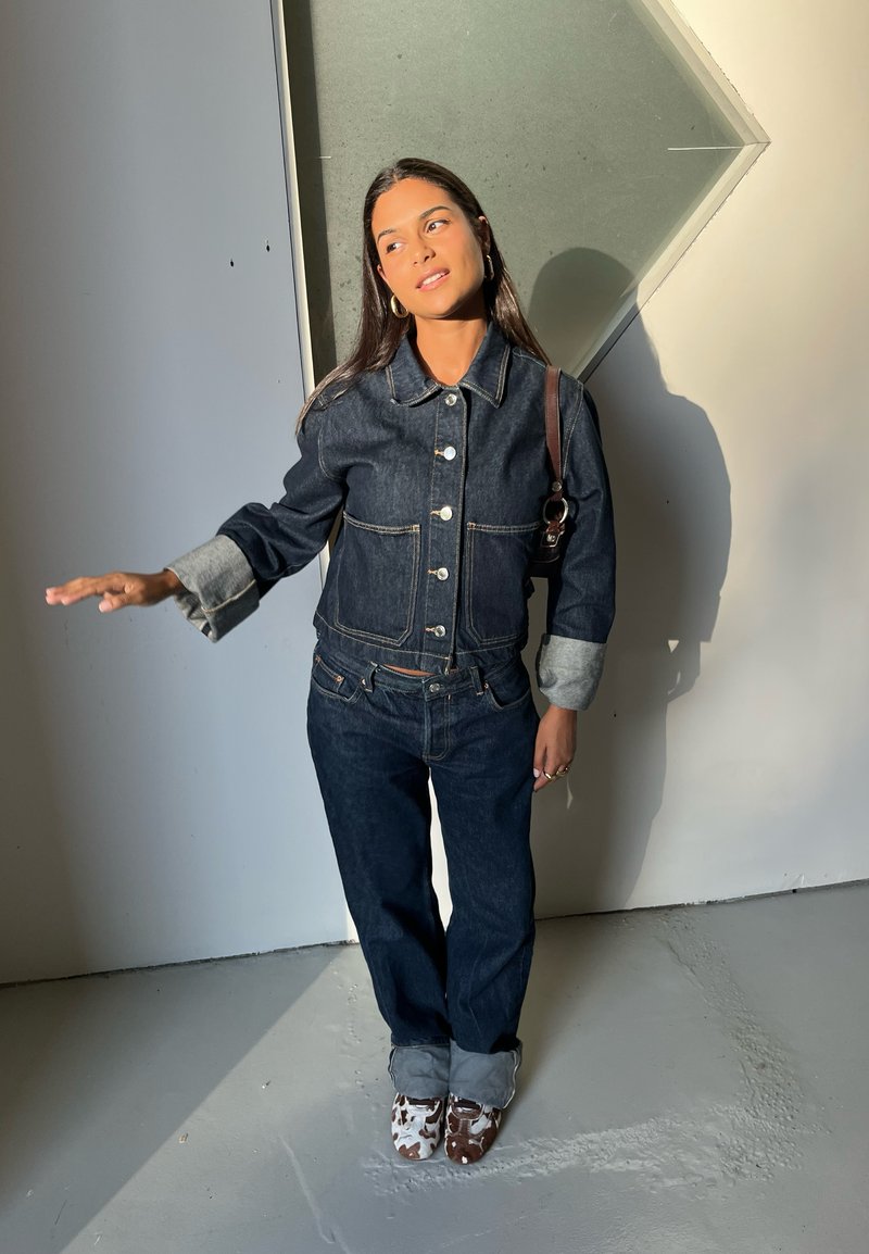 Woman in dark denim jacket and jeans with rolled cuffs, standing indoors near angled wall, looking right with left arm extended.