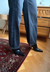Personne portant un jean large sombre et des bottines noires à talons aiguilles pointues, debout sur un tapis rouge à motifs et un sol en bois.