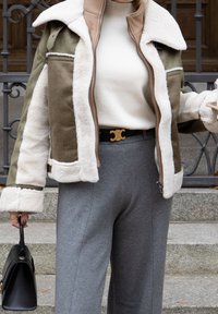 Jaqueta de shearling verde-azeitona e creme com um interior castanho, apresentando um fecho de correr na frente e um colarinho largo. Combinada com calças cinzas e uma bolsa preta.