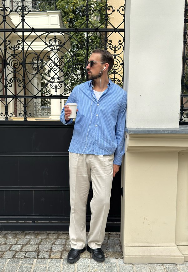 Hombre con gafas de sol, camisa azul a rayas, pantalones blancos y zapatos negros, sosteniendo una taza y de pie junto a una puerta metálica negra y un pilar beige.