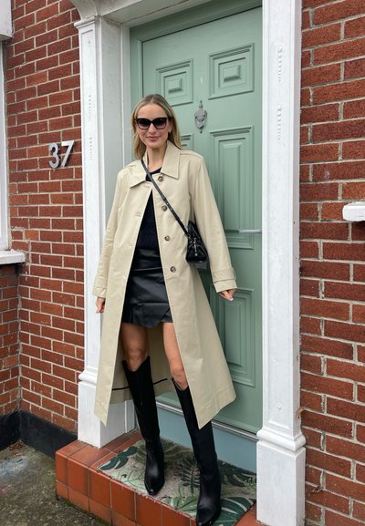 Gabardina beige, falda de cuero negra, botas de caña alta negras, bolso estrecho, gafas de sol, de pie junto a una puerta verde y una pared de ladrillo rojo.