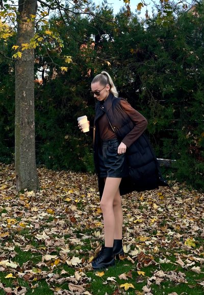 Chaleco de plumas negro sobre una camiseta de manga larga marrón, combinado con pantalones cortos de cuero negros y botas de tobillo. Sosteniendo una taza de café, rodeado de hojas de otoño.