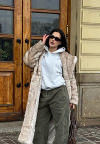 Manteau long en fourrure marron clair porté sur un sweat à capuche gris, associé à un pantalon large kaki. Lunettes de soleil noires et sac à main marron complètent la tenue.