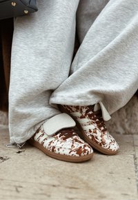 Personne portant un pantalon de survêtement gris et des baskets blanches avec imprimé vache marron et lacets marron, debout sur un sol carrelé près d'un sac à main noir.