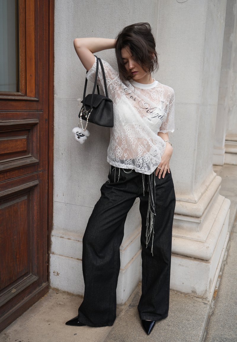 Mujer posando contra una pared de piedra con una blusa blanca de encaje transparente, pantalones negros de pierna ancha con cadenas colgantes, zapatos negros puntiagudos y sosteniendo un bolso negro.