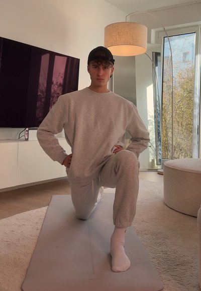 Joven con sudadera gris y pantalones arrodillado en una esterilla de yoga en el interior, cerca de una ventana, un televisor y una lámpara de pie con luz natural.