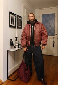 Tumši sarkana bumbvedēja jaka pār melnu kapuci, kopā ar platu tumšu džinsu biksēm. Rokās sarkankrāsas soma. Iekštelpu vide ar dekoratīviem elementiem.