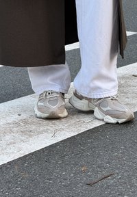 Baskets beige clair avec tissu en maille, accents texturés et semelle rembourrée. Associées à un pantalon blanc légèrement évasé et un manteau foncé.