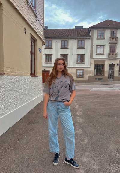 Camiseta gris de gran tamaño con diseño gráfico, jeans de pierna ancha de color azul claro y zapatillas negras; entorno urbano al aire libre con edificios al fondo.