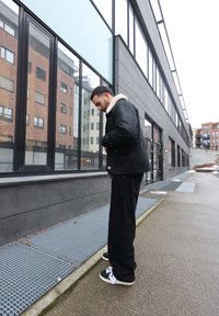 Homme en veste noire et pantalon debout devant un bâtiment moderne aux grandes fenêtres, ajustant le col de sa veste sur un trottoir gris.