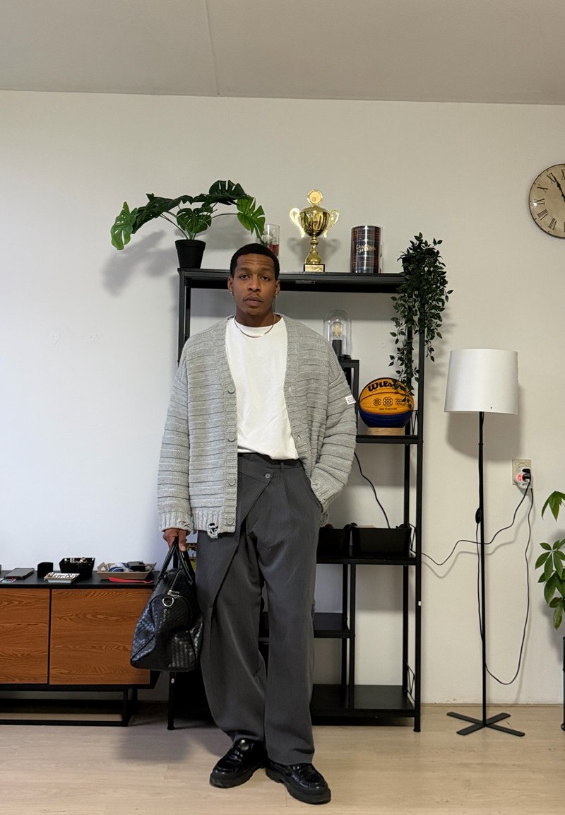 Homme en cardigan gris, chemise blanche et pantalon sombre tenant un sac noir, debout devant une étagère avec des plantes, des trophées et un ballon de basket.