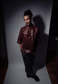 Chemise en cuir bordeaux à manches courtes avec un col, associée à un pantalon charbon et des chaussures noires, le tout sur un fond neutre.