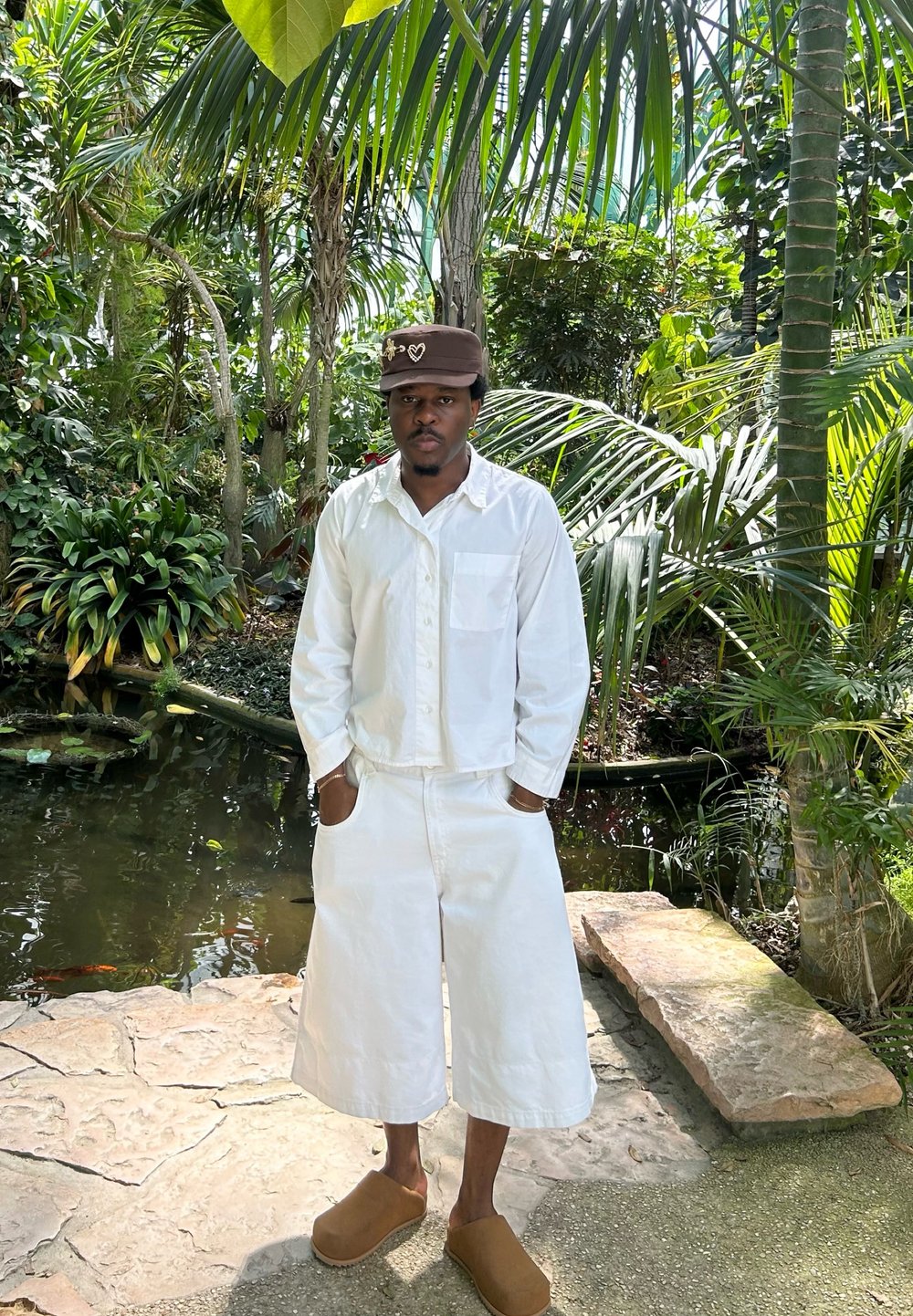 Witte, lichte katoenen shirt met een zak, gecombineerd met witte shorts tot de knie. Bruine suède klompen. Omringd door weelderig groen loof.