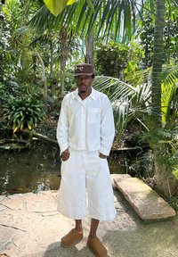 Witte, lichte katoenen shirt met een zak, gecombineerd met witte shorts tot de knie. Bruine suède klompen. Omringd door weelderig groen loof.