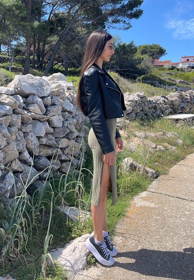 Chaqueta recortada de cuero negro, vestido midi de punto verde olivo con abertura lateral y zapatillas de plataforma grises sobre un camino de piedra junto a la vegetación.