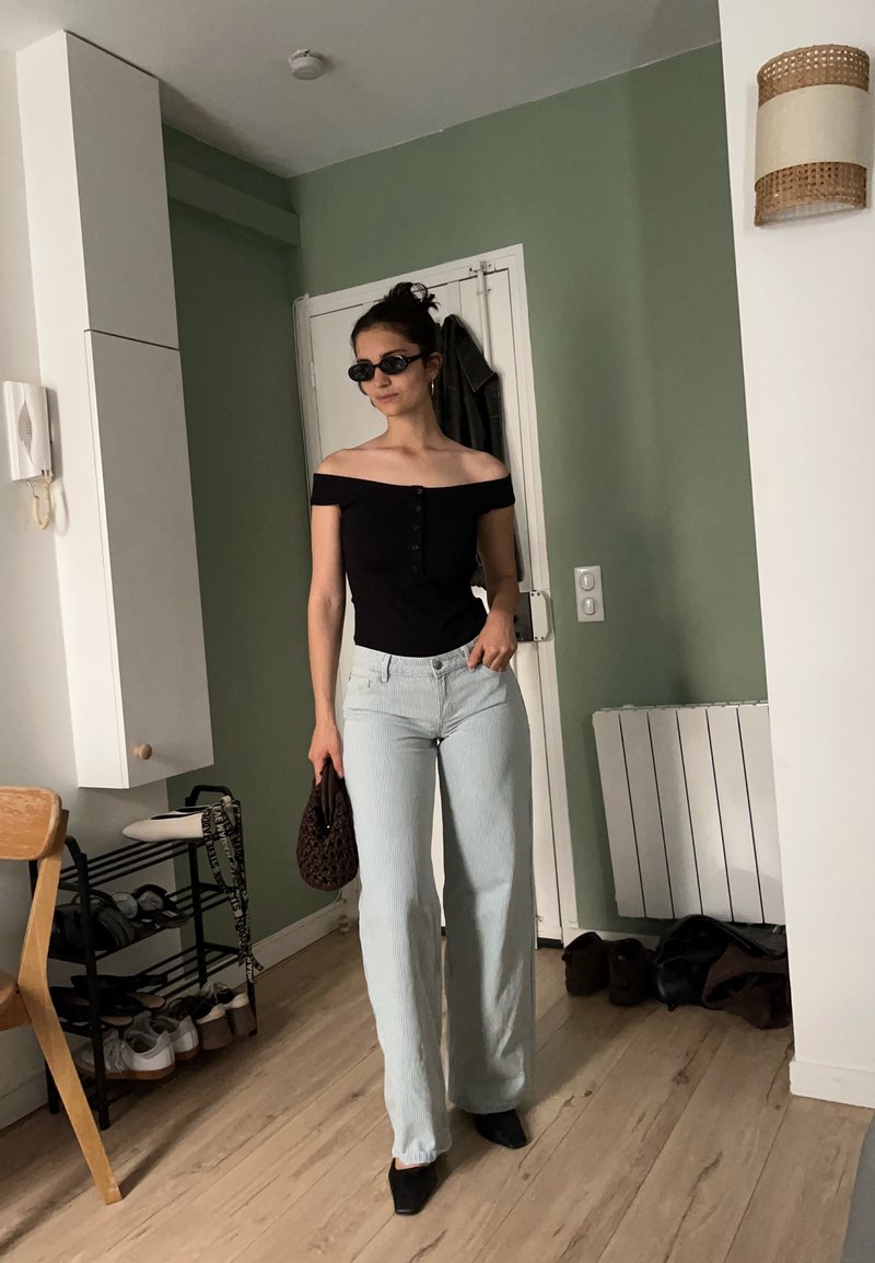 Femme portant un haut noir épaules dénudées, pantalon large bleu clair, chaussures noires, lunettes de soleil, tenant un sac marron, debout dans une pièce avec un mur vert et un sol en bois.