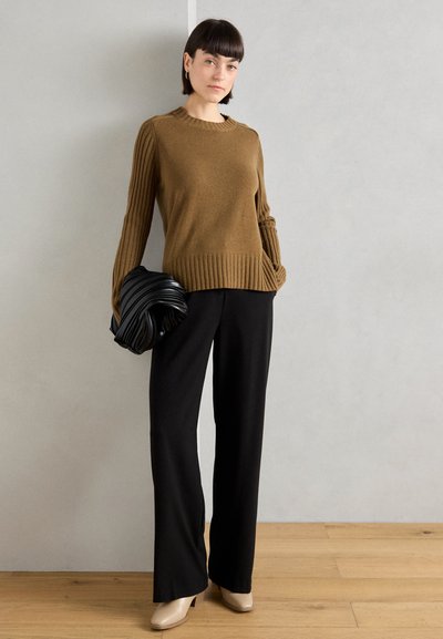 Suéter de punto texturizado marrón con mangas acanaladas, combinado con pantalones de pierna ancha negros y zapatos de tacón beige. Sosteniendo un bolso plisado negro.