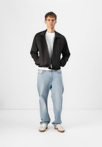 Svart zip-up jakke over en hvit t-skjorte, kombinert med lyseblå jeans og hvite sneakers. Jakken har krage og en glatt tekstur.