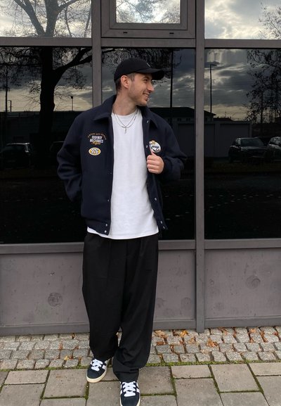 Chaqueta azul marino con parches y bordados, camiseta blanca, pantalones sueltos negros y zapatillas azules marinas con cordones blancos, de pie frente a un fondo de cristal.