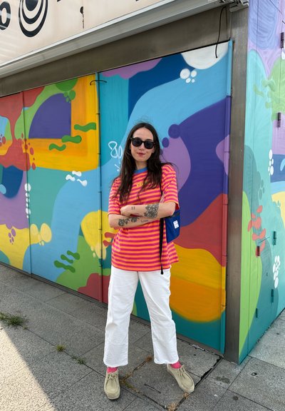 Camiseta a rayas rosa y naranja, pantalones anchos blancos, zapatos beige, gafas de sol negras, tatuajes visibles en los brazos, de pie frente a un mural colorido.