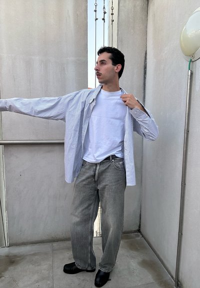 Hombre de pie en el balcón con camiseta blanca, camisa abierta a rayas, vaqueros grises y zapatos negros, sujetando el cuello de la camisa con una mano.